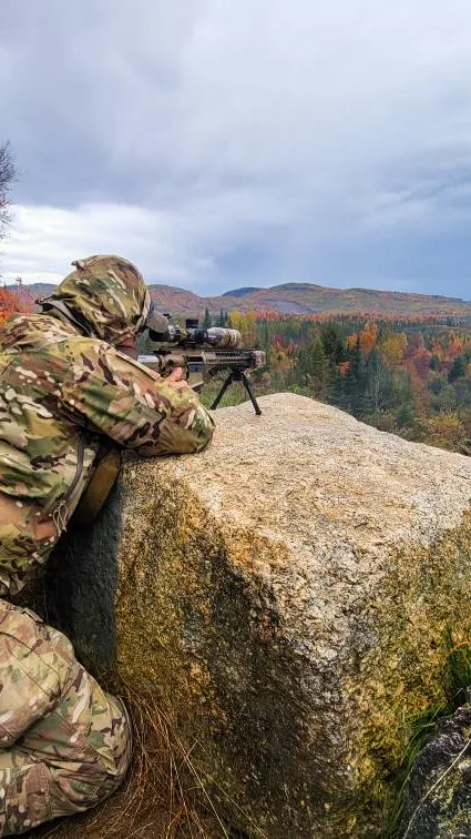 Shooter in camouflage aiming a rifle over a boulder toward autumn foliage and distant mountains