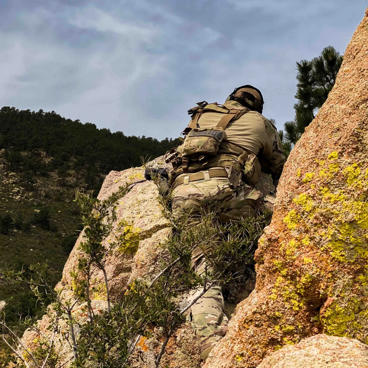 Shooter in tactical gear climbing a rock outcrop on a wooded mountainside