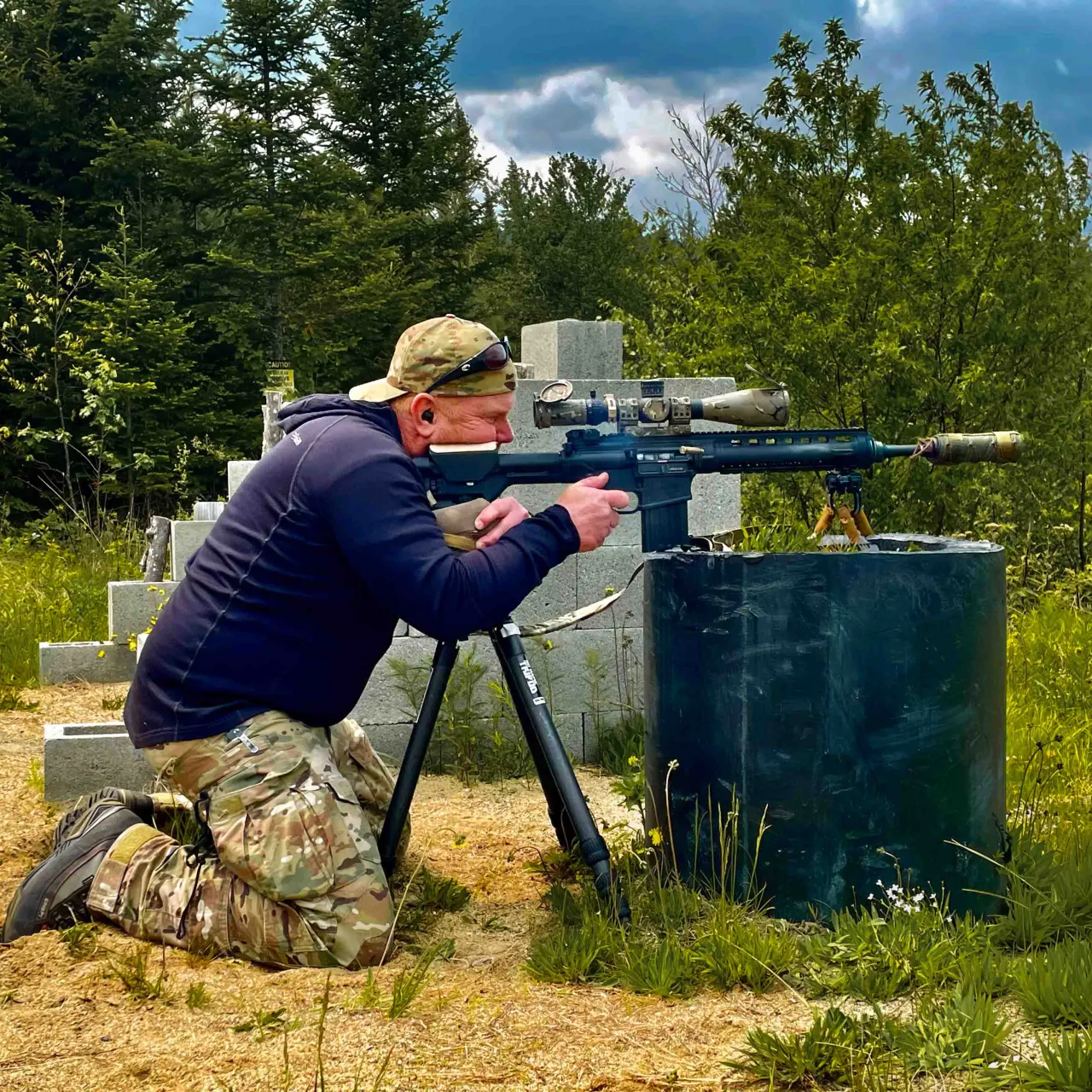 Student kneeling behind a rifle on a tripod, aiming over a concrete block barricade