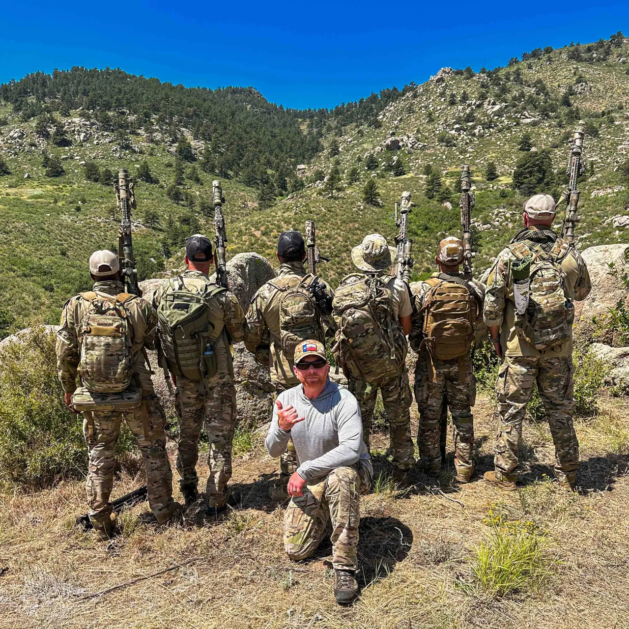 Class group standing on a mountain hillside with rifles on their packs, instructor kneeling in front