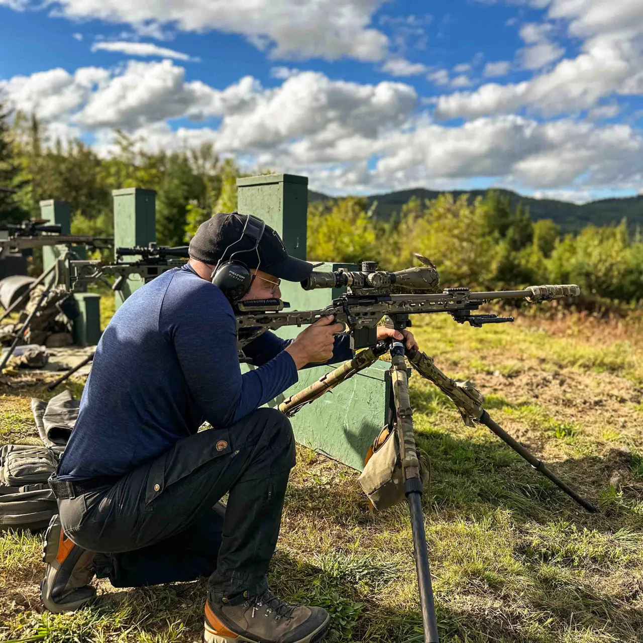 Shooter kneeling behind a rifle on a tripod at a barricade stage on the range