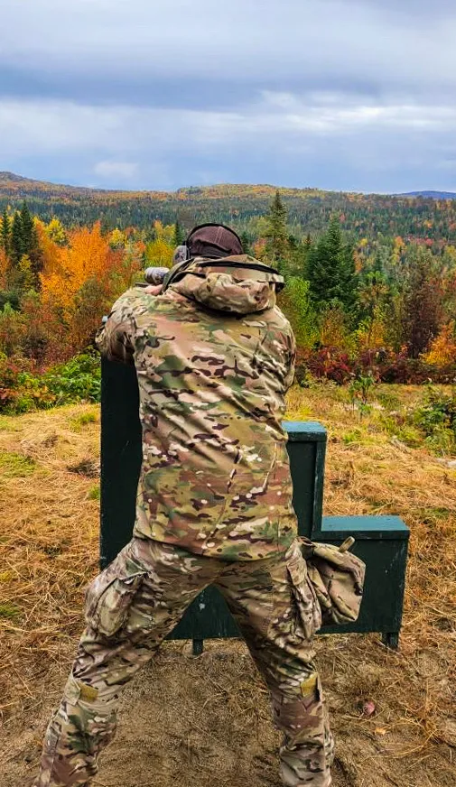 Shooter in camouflage aiming from a green barricade toward autumn hills
