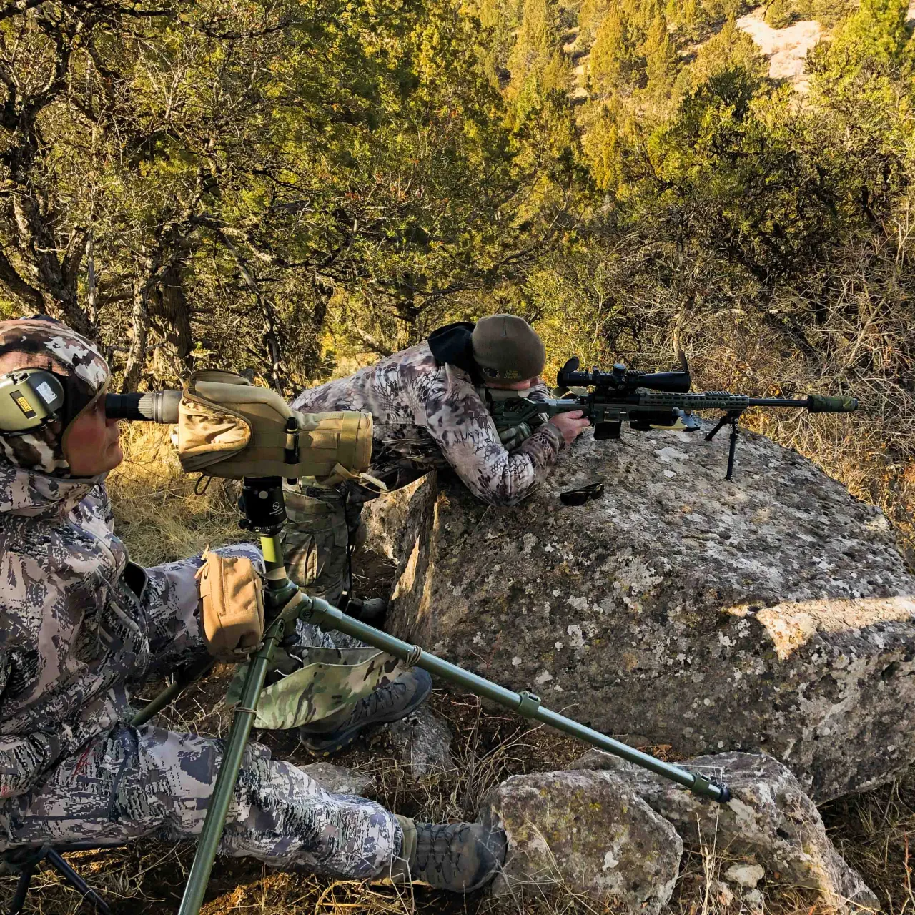 Spotter at a spotting scope beside a prone shooter on a rocky, wooded slope