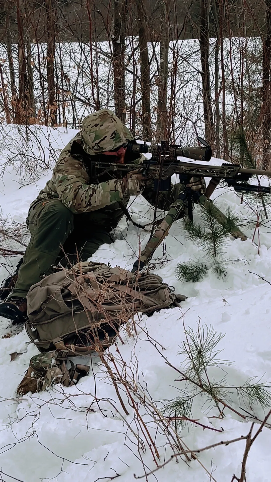 Shooter kneeling in the snow behind a rifle among pine trees